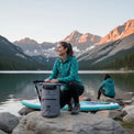 Woman kneeling by a lake with a grey RIXVA 15L waterproof insulated cooler bag, mountain landscape background.
